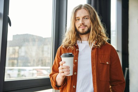 gutaussehender und cooler Geschäftsmann mit Pappbecher mit Kaffee zum Mitnehmen, während er am Fenster steht und im Büro in die Kamera schaut, lange blonde Haare, trendiges Hemd, Hipster-Stil
