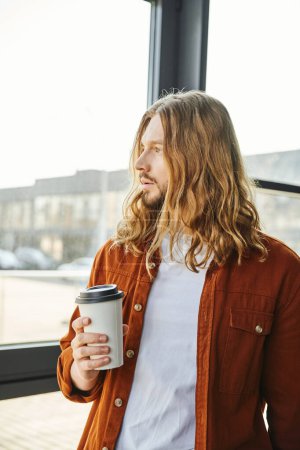 entrepreneur rêveur, aux cheveux longs et barbu dans des vêtements décontractés tenant tasse en papier avec café, debout près de la fenêtre et regardant loin, cool homme de style hipster dans le bureau moderne