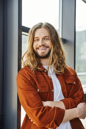 optimistic and stylish businessman with radiant smile looking at camera in office, handsome, hipster style, bearded, long haired, trendy shirt, successful and career oriented man