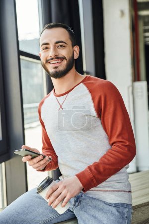 self-assured and stylish bearded man in casual clothes smiling at camera while holding mobile phone and paper cup with takeaway drink in contemporary office, young businessman 