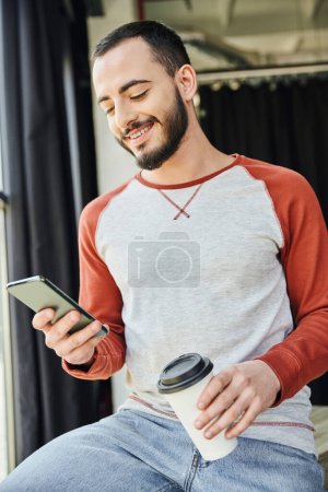 Photo for Charismatic entrepreneur, bearded and stylish, holding coffee to go in paper cup and browsing internet on smartphone, young and career oriented businessman in modern office - Royalty Free Image