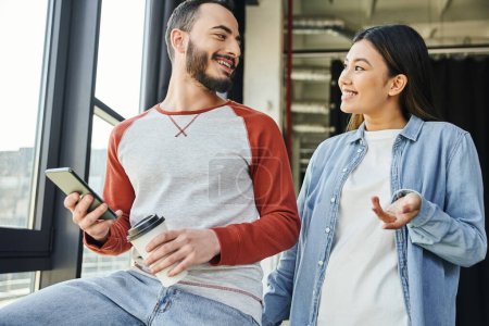 happy asian woman in stylish casual clothes talking to bearded colleague holding smartphone and paper cup with takeaway drink in modern office, young interracial entrepreneurs planning startup