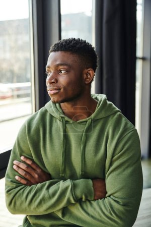 pensive african american businessman in green hoodie standing with folded arms and looking away near window in modern office, young entrepreneur 
