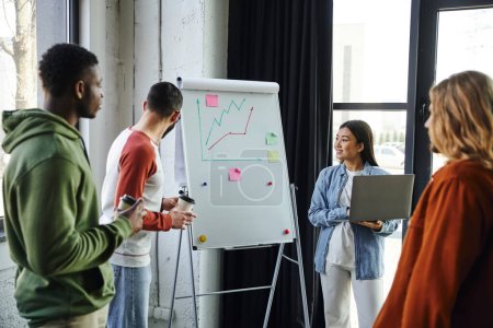 smiling asian woman with laptop and multiethnic men with coffee to go in paper cups looking at flip chart with graphs, young and creative team planning startup project in modern office