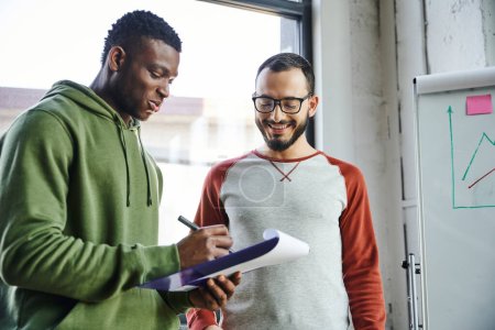 young african american businessman in green hoodie writing on clipboard near smiling bearded colleague in eyeglasses and flip chart with graphs in modern office, creative youth 
