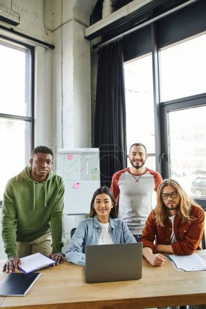 diverse group of optimistic, young and multicultural business people looking at camera near laptop, documents and flip chart with graphs on background in modern office space, creative collaboration