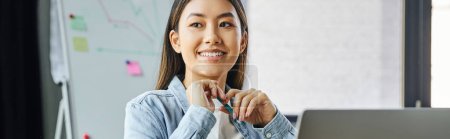 overjoyed asian woman with brunette hair and happy smile holding pen and looking away while sitting in modern office, young successful businesswoman, banner
