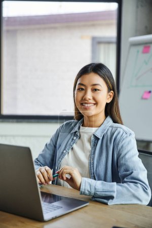 glückliche asiatische Geschäftsfrau mit brünetten Haaren und strahlendem Lächeln hält Stift in der Hand und blickt in die Kamera, während sie am Arbeitsplatz in der Nähe von Laptop im modernen Büro sitzt 