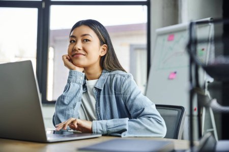 freudige asiatische Geschäftsfrau in blauem Jeanshemd, mit brünetten Haaren neben Laptop sitzend, die Hand vor dem Gesicht haltend und im Büro wegschauend, junge erfolgreiche Unternehmerin 