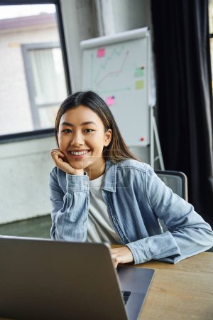 young and pleased asian businesswoman with brunette hair, in blue denim shirt looking at camera near laptop and flip chart with graphs on blurred background in contemporary office