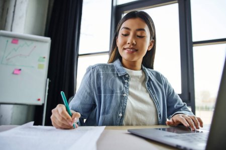 lächelnde Asiatin mit brünetten Haaren, in blauem Jeanshemd, das am Laptop arbeitet und auf Dokumente in der Nähe von Flipchart auf verschwommenem Hintergrund in zeitgenössischen Büros schreibt, erfolgreiches Unternehmertum