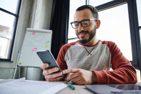 joyful and bearded businessman in stylish eyeglasses networking on smartphone while sitting next to laptop, documents and flip chart on blurred background in modern office
