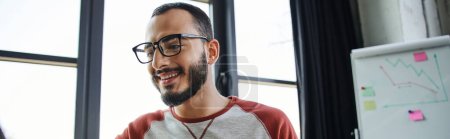 portrait of young and happy bearded manager in eyeglasses working in modern office near flip chart with business analytics on blurred background, business lifestyle, banner