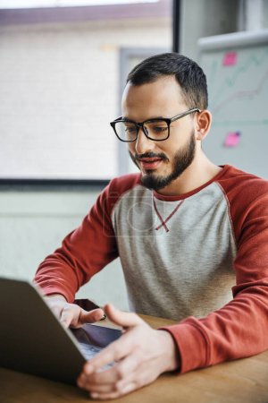 positive and concentrated bearded entrepreneur in eyeglasses and casual clothes sitting at workplace and working on laptop on blurred foreground in modern office, successful youth