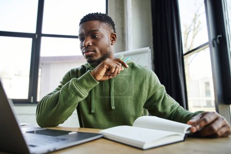 young and handsome african american businessman in green stylish hoodie looking at laptop near notebook while sitting at workplace and working on blurred foreground in modern office