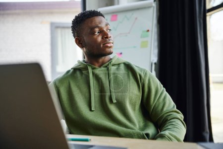 Téléchargez les photos : Jeune entrepreneur afro-américain en sweat à capuche vert assis et regardant loin tout en planifiant démarrage près de flip chart avec l'analyse des affaires dans le bureau moderne, le développement professionnel - en image libre de droit