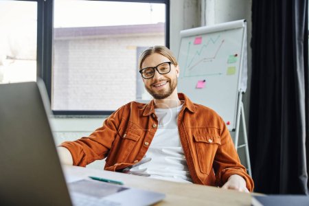 glücklicher und optimistischer bärtiger Geschäftsmann in Brille und schickem Hemd sitzt am Laptop und blickt neben Flipchart auf verschwommenem Hintergrund in zeitgenössisches Büro