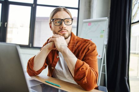 stylish and pensive businessman in eyeglasses and trendy casual clothes looking away near laptop and pen on work desk next to flip chart with graphs on blurred background in modern office