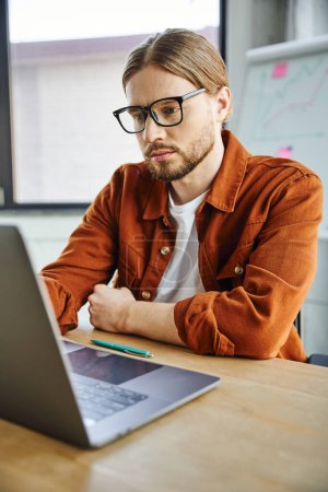 Aufmerksame und konzentrierte bärtige Unternehmerin in trendiger Freizeitkleidung und Brille, die am Laptop neben dem Stift auf dem Schreibtisch im modernen Büro arbeitet, Business Lifestyle