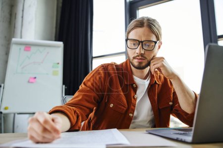 hombre de negocios barbudo serio en camisa de moda y anteojos mirando el documento mientras está sentado cerca de la computadora portátil y el rotafolio con gráficos sobre fondo borroso en la oficina