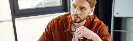 portrait of thoughtful and bearded entrepreneur in trendy casual shirt holding eyeglasses and looking away while working in contemporary office, business lifestyle, banner