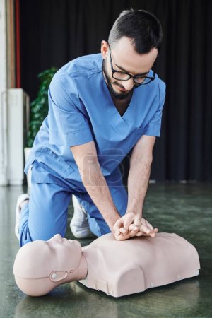 junger medizinischer Instruktor mit Brille und Uniform beim Durchführen von Brustkompressionen an der CPR-Schaufensterpuppe auf dem Boden im Schulungsraum, effektive lebensrettende Fähigkeiten und Notfallvorsorgekonzept
