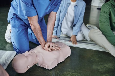 cardiopulmonary resuscitation, partial view of medical instructor doing chest compressions on CPR manikin near young participants of first aid seminar, life-saving skills and techniques concept