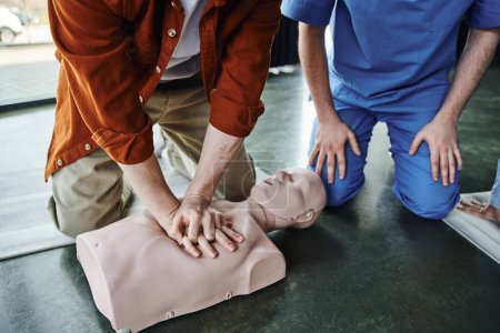 Teilansicht eines jungen Mannes, der bei einem Erste-Hilfe-Seminar in der Nähe von professionellen Rettungssanitätern Brustkompressionen an CPR-Schaufensterpuppen durchführt, Herz-Lungen-Wiederbelebung, lebensrettende Fähigkeiten und Techniken