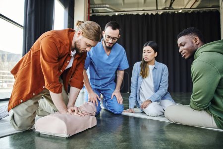 junges multiethnisches Team und professionelle Sanitäter, die den Afrikaner beim praktischen Erste-Hilfe-Lernen dabei beobachten, wie er Brustkompressionen an einer CPR-Schaufensterpuppe macht, effektives Konzept für lebensrettende Fähigkeiten