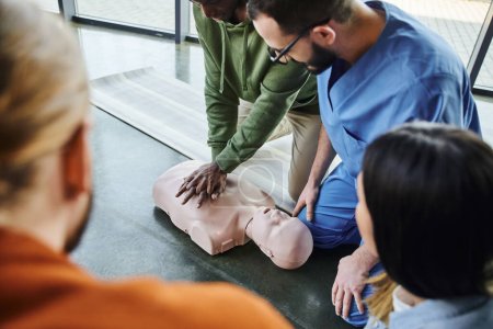 Afrikanisch-amerikanischer Mann übt Brustkompressionen und Herz-Lungen-Wiederbelebung an CPR-Schaufensterpuppe in der Nähe von Ausbildern und jungen Teilnehmern eines Erste-Hilfe-Seminars im Schulungsraum