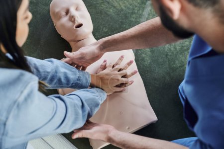 Ansicht von oben: junge Frau übt Brustkompressionen an CPR-Schaufensterpuppe während des praktischen Lernens auf einem Erste-Hilfe-Seminar in der Nähe eines medizinischen Instruktors, lebensrettende Fähigkeiten und Techniken
