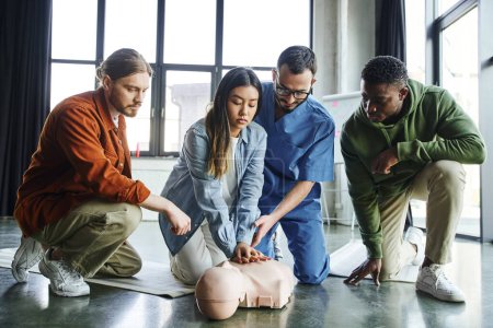 junge Asiatin macht Brustkompressionen an CPR-Schaufensterpuppen während sie Herz-Lungen-Wiederbelebung in der Nähe paramedizinischer und multiethnischer Teilnehmer praktiziert, lebensrettende Fähigkeiten und Techniken