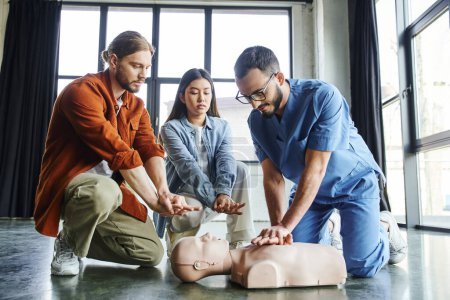professioneller Rettungssanitäter mit Brille und Uniform, der Brustkompressionen an der CPR-Schaufensterpuppe in der Nähe eines jungen Mannes und einer asiatischen Frau während eines Erste-Hilfe-Trainings zeigt, effektives lebensrettendes Kompetenzkonzept