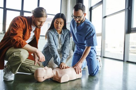 Asiatin übt Brustkompressionen an CPR-Schaufensterpuppe in der Nähe junger Mann und medizinischer Ausbilder in Brille und Uniform während eines Erste-Hilfe-Seminars, Notfallpräventionskonzept