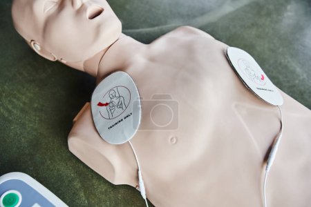 Hohe Blickwinkel auf Herz-Lungen-Wiederbelebungsübungspuppe mit Defibrillatorkissen auf dem Boden im Schulungsraum, medizinisches Gerät für Erste-Hilfe-Training und Kompetenzentwicklung