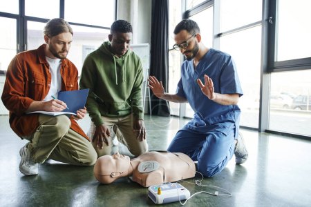 young paramedic explaining cardiac resuscitation techniques to interracial participants near CPR manikin with automated external defibrillator, effective life-saving skills and techniques concept