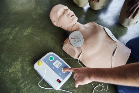 Teilansicht des professionellen medizinischen Instruktors, der Defibrillator an der CPR-Schaufensterpuppe bedient, in der Nähe der jungen Teilnehmer des Erste-Hilfe-Seminars, Blickwinkel, Gesundheitsfürsorge und lebensrettendes Technikkonzept