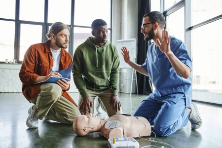 healthcare worker gesturing and talking next to CPR manikin with defibrillator near african american participant and young man writing in notebook, effective life-saving skills and techniques concept