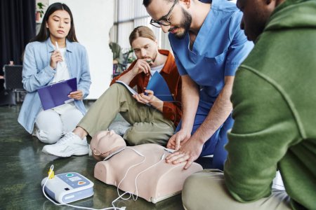 first aid seminar, medical instructor applying defibrillator pads on CPR manikin near multiethnic team with clipboard and notebook in training room, health care and life-saving techniques concept