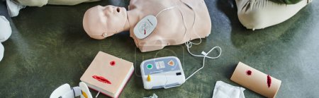 top view of CPR manikin, automated external defibrillator and wound care simulators near cropped participants of first aid training seminar, health care and emergency preparedness concept, banner
