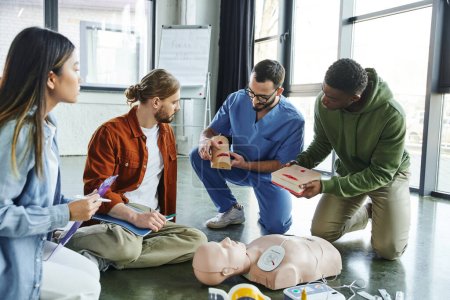 medical instructor and african american man holding wound care simulators near CPR manikin, defibrillator and multiethnic participants with notebook and clipboard, life-saving skills concept