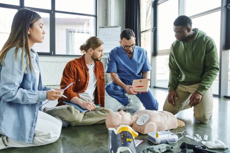 Un ambulancier paramédical professionnel présente un simulateur de soins des plaies à une équipe multiculturelle près d'un mannequin de RCR, d'un défibrillateur et d'un équipement médical dans une salle d'entraînement, un concept efficace de premiers soins et de compétences vitales