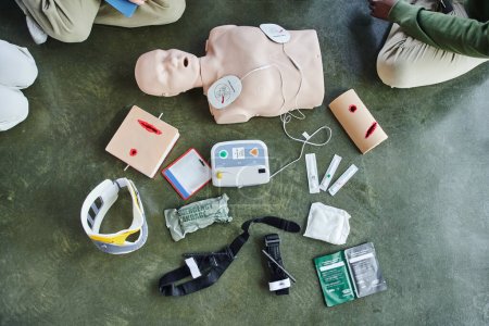 first aid seminar, cropped view of young people near CPR manikin, defibrillator, wound care simulators, bandages, compression tourniquets, syringes and neck brace in training room, top view