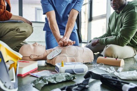 partial view of paramedic doing chest compressions on CPR manikin near multiethnic participants and medical equipment, defibrillator, wound care simulators, tourniquets, first aid training seminar
