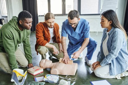 Gesundheitshelfer stampft Wunde mit Verband auf Simulator und zeigt multikulturellem Team in der Nähe von CPR-Schaufensterpuppe, Defibrillator und medizinischen Geräten lebensrettende Fähigkeiten, Notfallreaktionskonzept