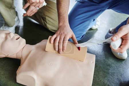 Hochwinkelaufnahme des medizinischen Instruktors beim Tamponieren einer Wunde am Simulator mit Verband in der Nähe von CPR-Schaufensterpuppe und jungem Mann während eines Erste-Hilfe-Trainings, Notfallkonzept, Ausschnitt