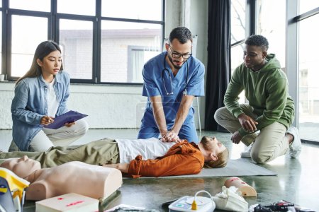 professionelle Sanitäter üben Brustkompressionen am Mann in der Nähe von CPR-Schaufensterpuppen, medizinischem Gerät und multiethnischen Teilnehmern eines Erste-Hilfe-Seminars, effektives Konzept zur Rettung von Leben