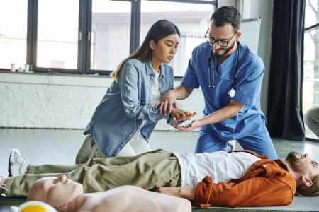 Ärztlicher Instruktor zeigt der jungen asiatischen Frau in Männernähe, die neben der CPR-Schaufensterpuppe im Schulungsraum liegt, die Position der Hände zur Herz-Lungen-Wiederbelebung, effektives Konzept der lebensrettenden Fähigkeiten und Techniken