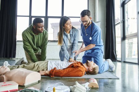 homme afro-américain regardant ambulancier aidant femme asiatique faire des compressions thoraciques sur participant couché près du mannequin de RCR et de l'équipement médical, compétences et techniques de sauvetage concept