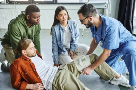 healthcare worker applying compressive bandage on leg of man near interracial participants during first aid seminar in training room, bleeding prevention techniques concept